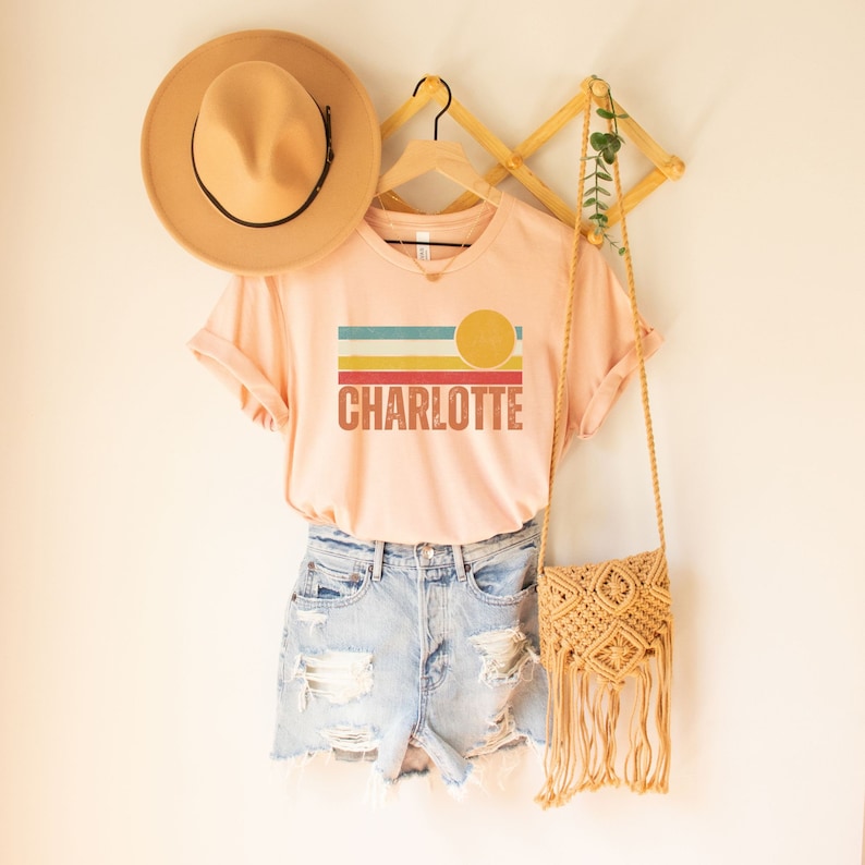 May include: A peach-colored t-shirt with a retro sunset graphic and the name "CHARLOTTE" printed in red. The shirt is hanging on a wooden hanger with a straw hat and a woven bag.