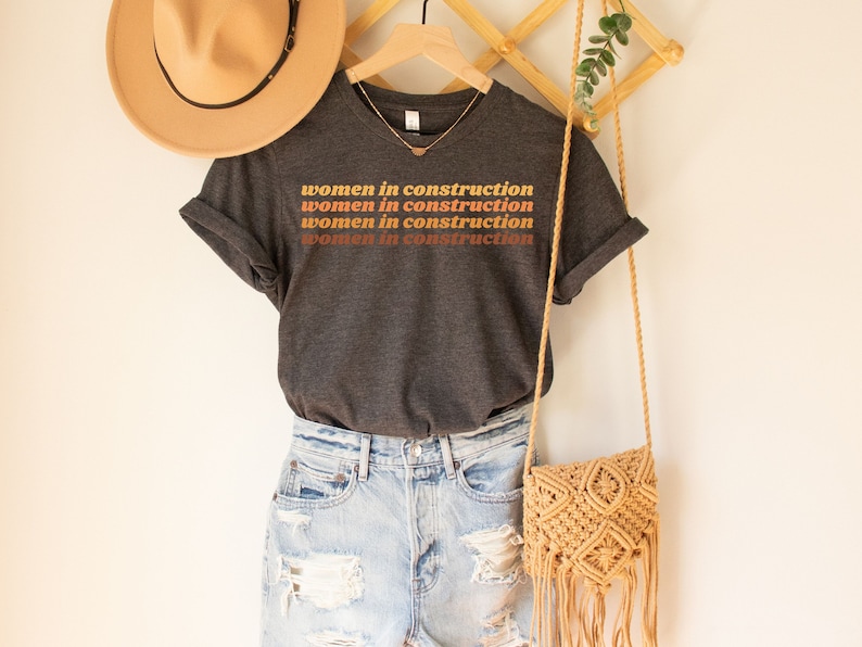 Puede incluir: Una camiseta gris oscura con el texto "women in construction" repetido cuatro veces en un degradado de naranja, marr&oacute;n y amarillo. La camiseta est&aacute; colgada en un estante de madera con un sombrero marr&oacute;n y un bolso tejido.