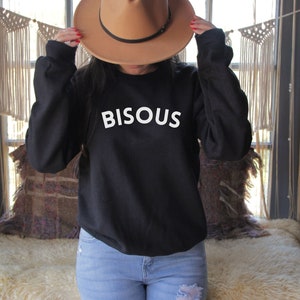 May include: A black sweatshirt with the word "BISOUS" in white block letters. A tan fedora-style hat with a black band is held up. The person is wearing light blue jeans.