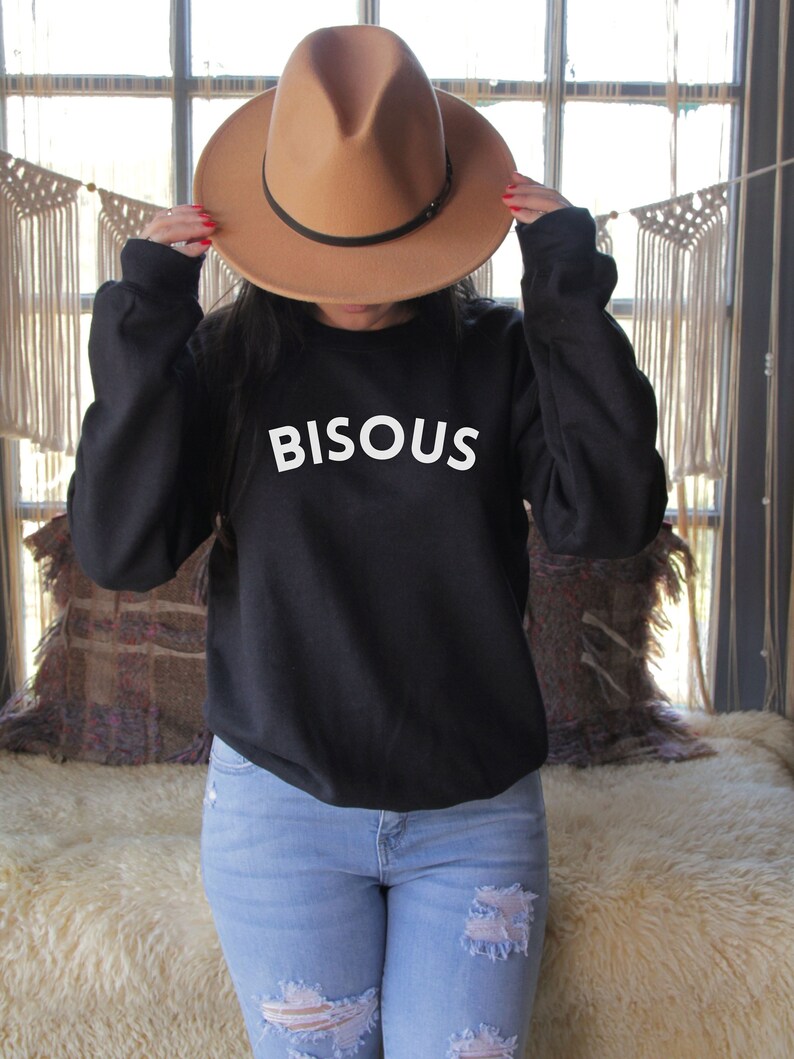 May include: A black sweatshirt with the word "BISOUS" in white block letters. A tan fedora-style hat with a black band is held up. The person is wearing light blue jeans.