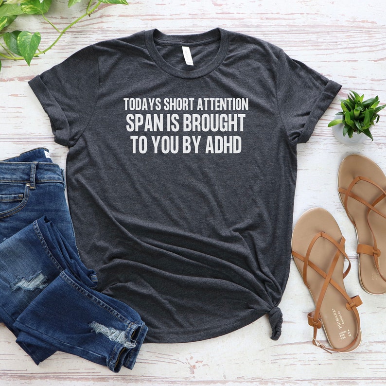 K&ouml;nnte beinhalten: Ein dunkelgraues T-Shirt mit wei&szlig;em Text, der "TODAYS SHORT ATTENTION SPAN IS BROUGHT TO YOU BY ADHD" lautet.
