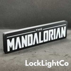 May include: Black and white sign with the text "THE MANDALORIAN" in white lettering on a black background. The sign is made of a hard material and has a slightly raised edge.