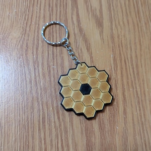 May include: A gold and black keychain featuring a honeycomb design with a central black hexagon. The keychain has a silver key ring and chain, and is likely a novelty item or souvenir.