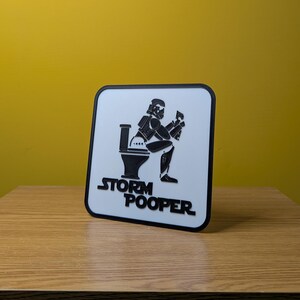 May include: White sign with a black outline and a black and white illustration of a Stormtrooper sitting on a toilet. The text "Storm Pooper" is printed below the illustration.