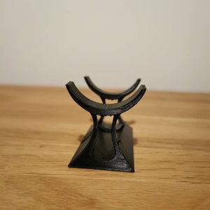 May include: A black, 3D-printed stand with a curved top and a rectangular base. The stand is designed to hold an item, with two curved cradles. The stand is made of a textured material and sits on a wooden surface.