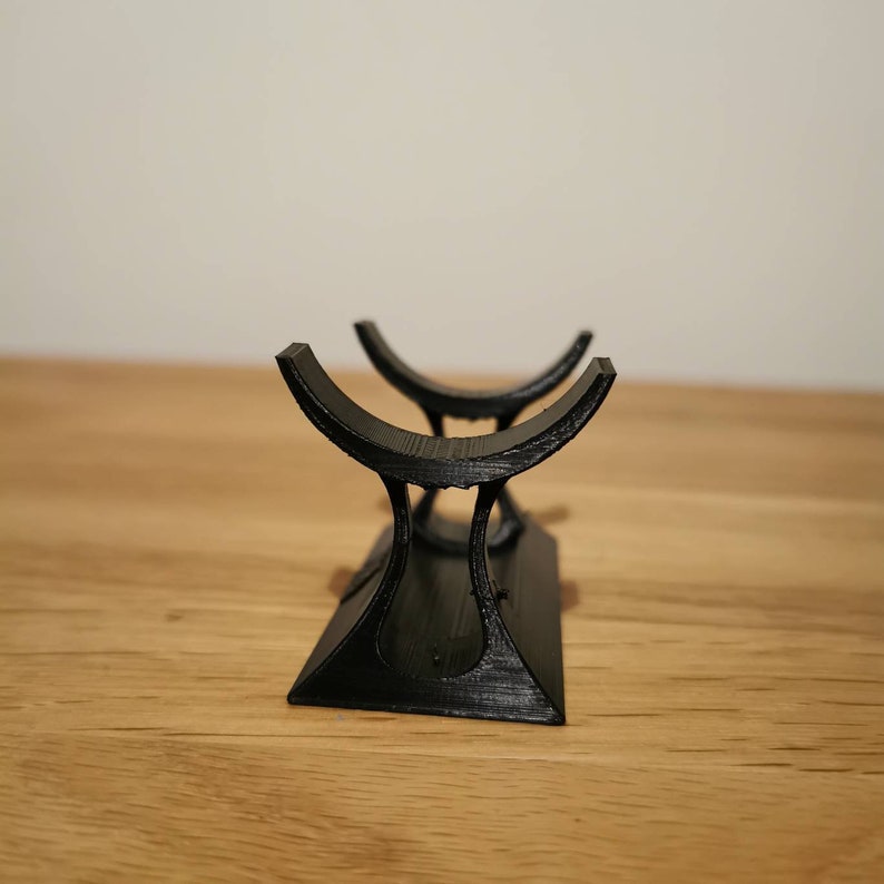 May include: A black, 3D-printed stand with a curved top and a rectangular base. The stand is designed to hold an item, with two curved cradles. The stand is made of a textured material and sits on a wooden surface.