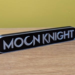 Puede incluir: Un letrero negro y plateado con el texto "MOON KNIGHT" en una fuente estilizada.