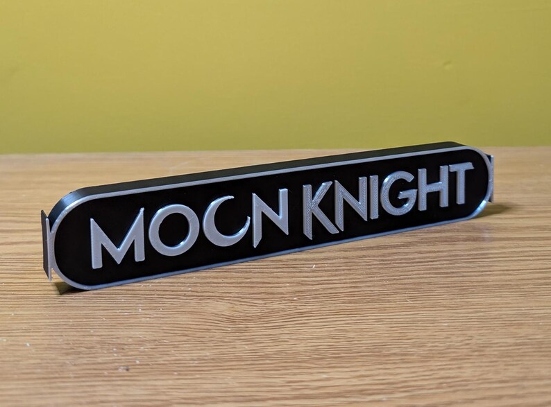 Puede incluir: Un letrero negro y plateado con el texto "MOON KNIGHT" en una fuente estilizada.