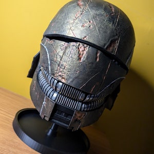 May include: A weathered, dark gray and brown helmet with a black visor and a detailed, textured surface. The helmet is on a black circular stand.