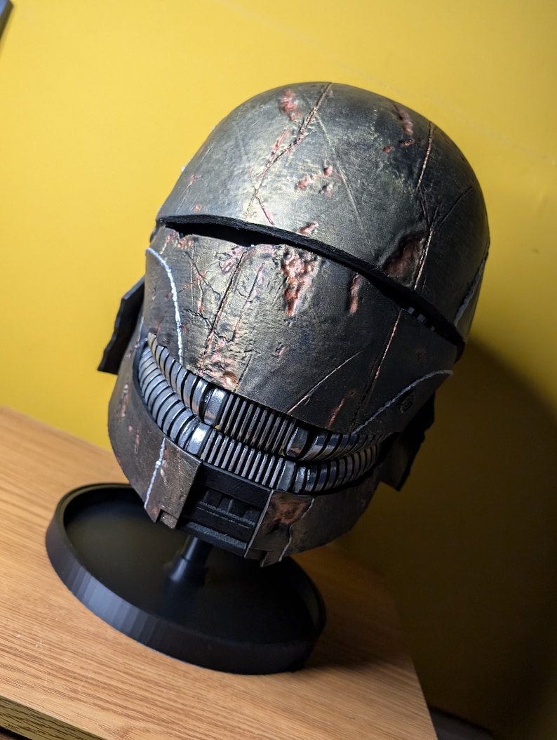 May include: A weathered, dark gray and brown helmet with a black visor and a detailed, textured surface. The helmet is on a black circular stand.