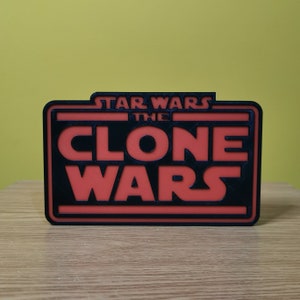 以下が含まれることがあります： 「スター・ウォーズ クローン・ウォーズ」の文字が書かれた黒と赤の3Dプリントされた看板です。