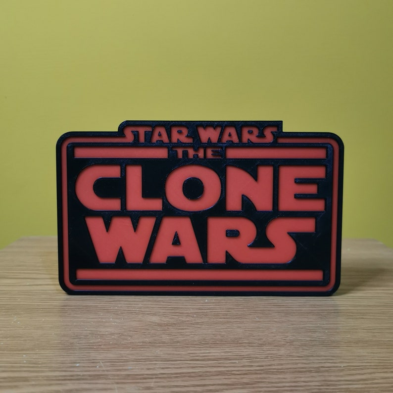 以下が含まれることがあります： 「スター・ウォーズ クローン・ウォーズ」の文字が書かれた黒と赤の3Dプリントされた看板です。