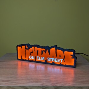 May include: Illuminated sign with the text "A Nightmare on Elm Street" in orange and white. The sign has a black outline and is lit from within, creating a glowing effect. It is placed on a wooden surface.
