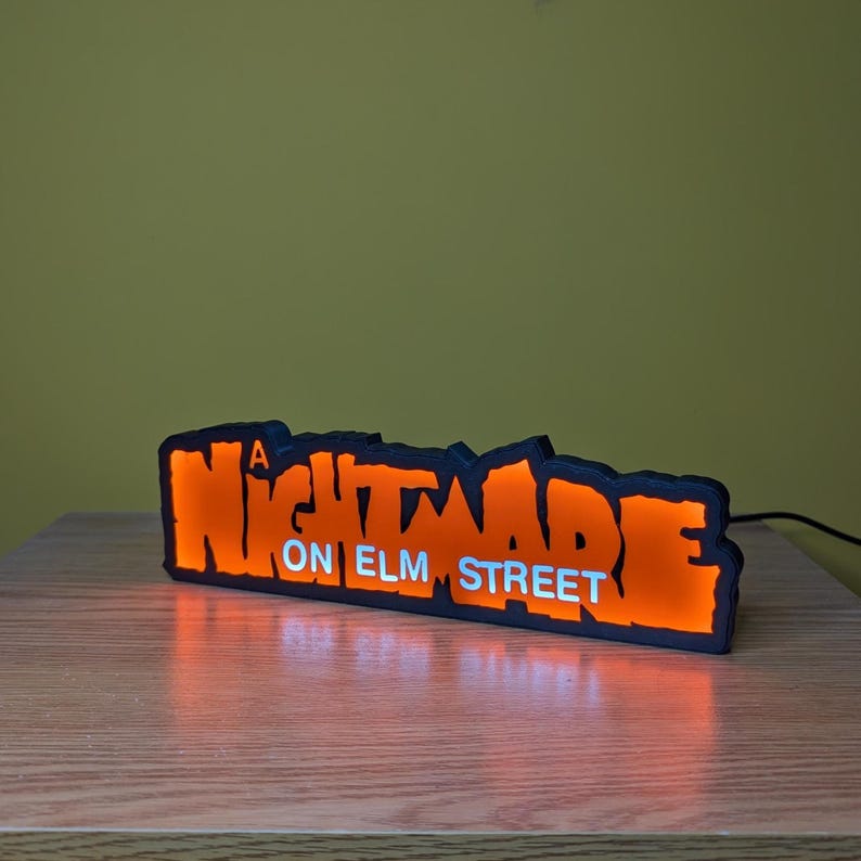 May include: Illuminated sign with the text "A Nightmare on Elm Street" in orange and white. The sign has a black outline and is lit from within, creating a glowing effect. It is placed on a wooden surface.