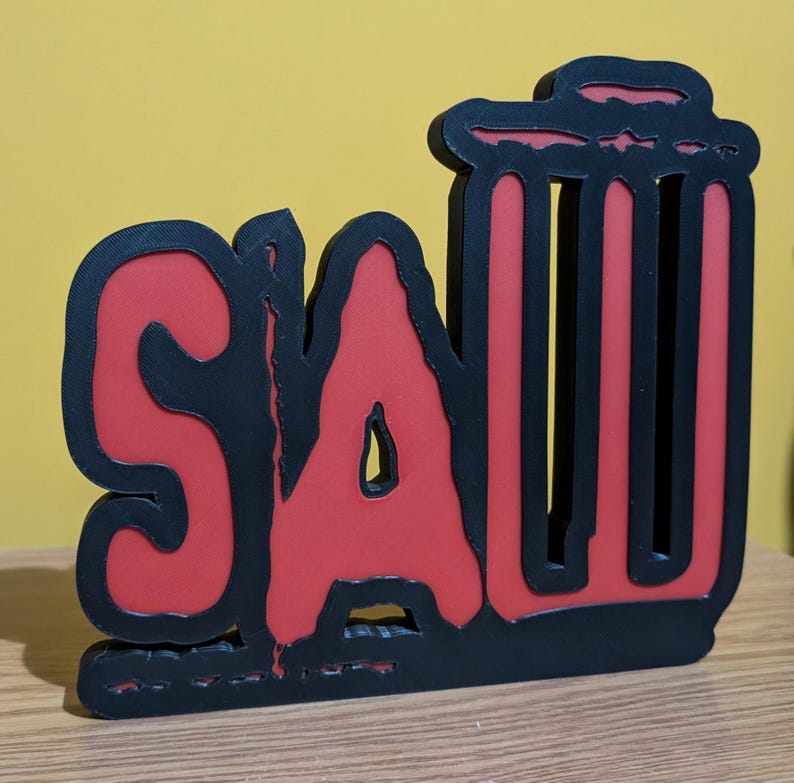 May include: Black and red 3D wall art with the word "SAW" in a stylized font. The "A" is a large, bold letter with a jagged edge. The "W" is a stylized representation of a trash can.