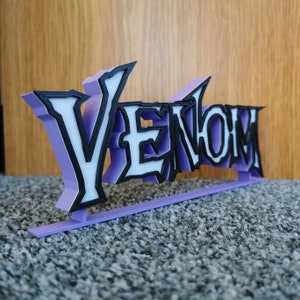 May include: A 3D-printed sign reading "Venom". The letters are white with black outlines and a light purple backing. The sign is mounted on a light purple base. The sign is likely a decorative item.