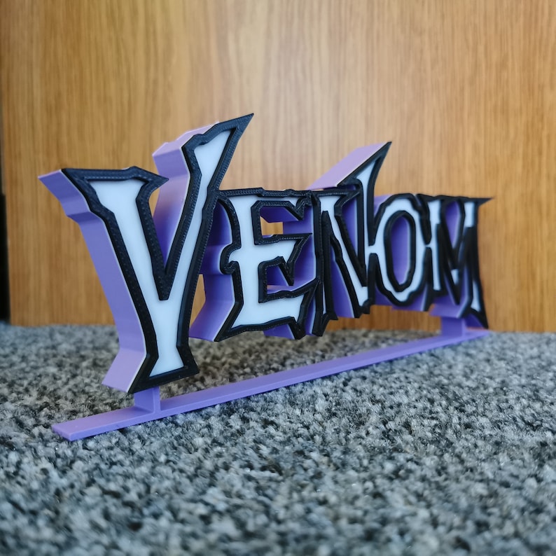 May include: A 3D-printed sign reading "Venom". The letters are white with black outlines and a light purple backing. The sign is mounted on a light purple base. The sign is likely a decorative item.