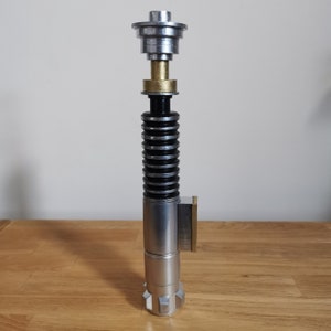 May include: A silver and black lightsaber prop with a gold accent. The lightsaber is made of metal and has a detailed design.