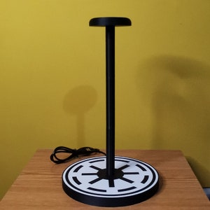 Könnte beinhalten: Schwarzer Kopfhörerständer mit einem runden Sockel, der das Symbol der Rebellenallianz aus Star Wars in Weiß und Schwarz zeigt. Der Ständer hat eine schwarze Metallstange und eine runde Oberseite.