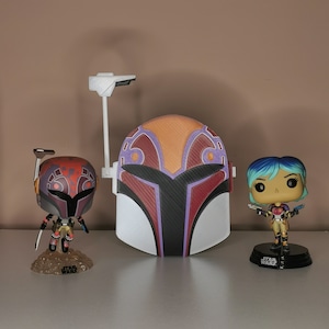 Może przedstawiać: Trzy figurki Star Wars, w tym hełm Mandalorianina, postać z czerwono-czarnym hełmem i postać kobieca z niebieskimi włosami. Wszystkie figurki znajdują się na białej powierzchni.