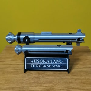 May include: A silver lightsaber prop with a black handle and a silver stand. The stand has a black plaque with the text "Ahsoka Tano The Clone Wars".