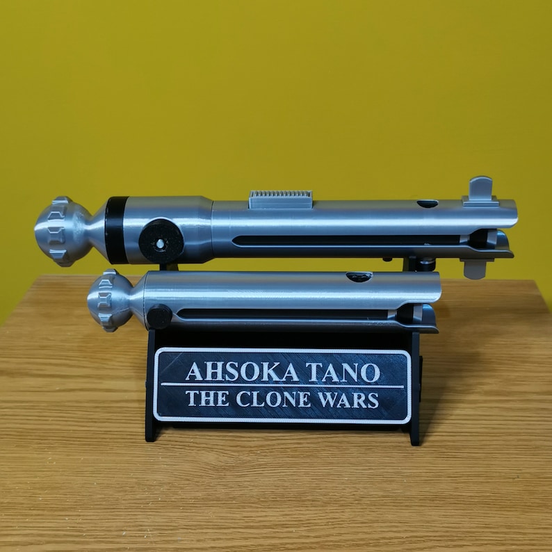May include: A silver lightsaber prop with a black handle and a silver stand. The stand has a black plaque with the text "Ahsoka Tano The Clone Wars".