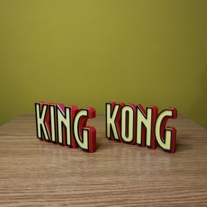 Pu&ograve; includere: Un cartello di parole in 3D di colore rosso e giallo che recita "KING KONG" con contorni neri. Il cartello &egrave; fatto di plastica e si trova su una superficie di legno.