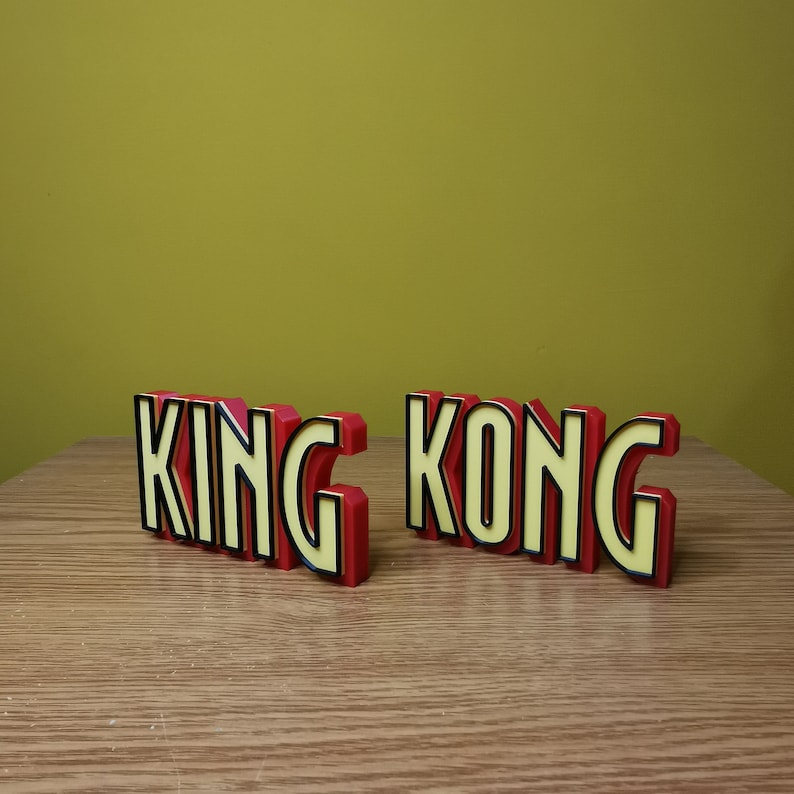 Pu&ograve; includere: Un cartello di parole in 3D di colore rosso e giallo che recita "KING KONG" con contorni neri. Il cartello &egrave; fatto di plastica e si trova su una superficie di legno.