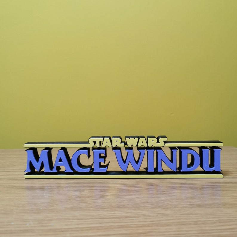 May include: A black and yellow 3D printed sign with the text "STAR WARS MACE WINDU" in blue letters.