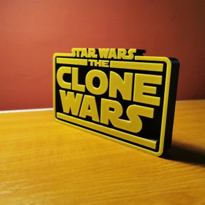 May include: A black and yellow 3D printed sign with the text "Star Wars The Clone Wars" in a bold, blocky font.