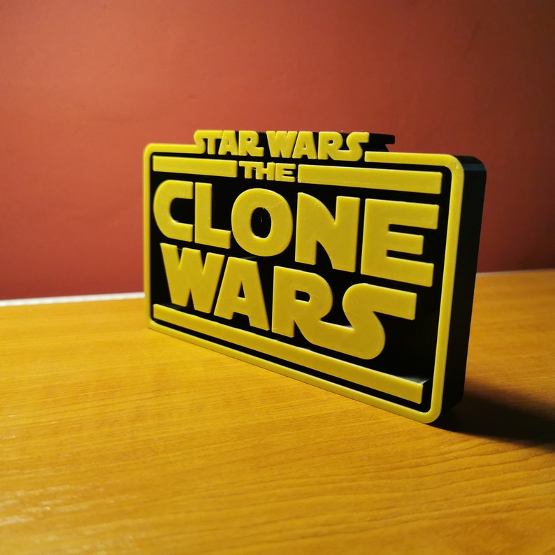 May include: A black and yellow 3D printed sign with the text "Star Wars The Clone Wars" in a bold, blocky font.