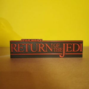 May include: A black 3D printed sign with the text "STAR WARS RETURN OF THE JEDI" in red.