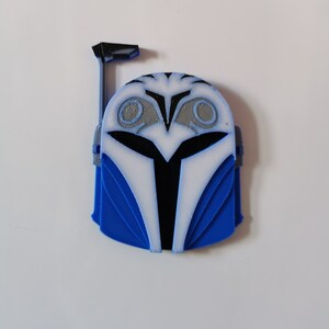 Op de afbeelding: Een blauwe en witte 3D-geprinte Mandalorian helm met een zwart vizier. De helm heeft een zilveren ontwerp aan de bovenkant en een zwarte antenne.