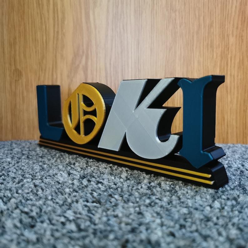 May include: A black, gold, silver, and teal 3D printed sign that reads "LOKI" with a gold circle in the center of the "O".