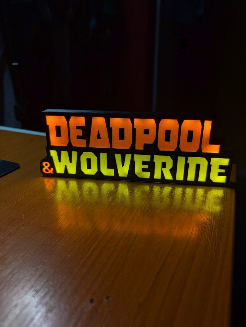 Pu&ograve; includere: Un'insegna rettangolare nera con le parole "DEADPOOL & WOLVERINE" in lettere illuminate arancioni e gialle. L'insegna &egrave; su una superficie di legno, con il riflesso del testo visibile.