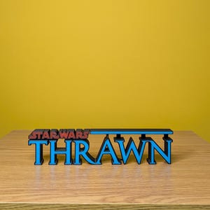 May include: A 3D printed sign with the words "STAR WARS" in red and "THRAWN" in blue. The sign is made of black plastic and has a blue stripe across the top.