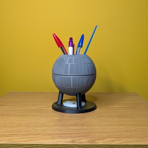May include: A gray Death Star-shaped desk organizer with a black base and three legs. It holds three pens, one red, one blue, and one purple.