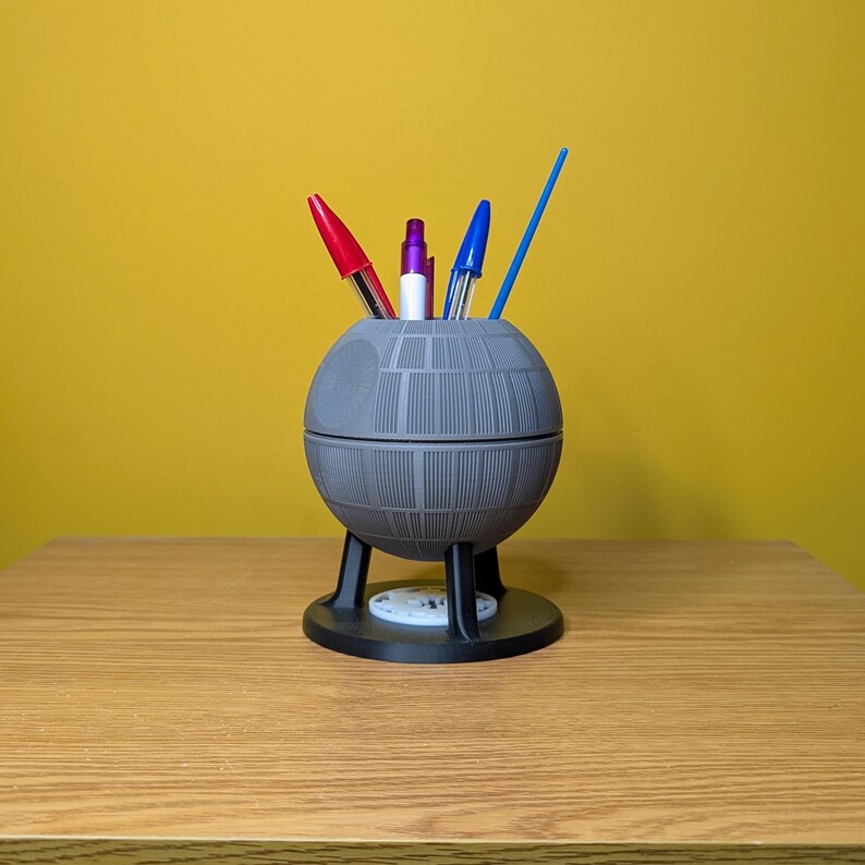 May include: A gray Death Star-shaped desk organizer with a black base and three legs. It holds three pens, one red, one blue, and one purple.