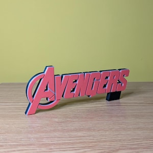 May include: A red and black Avengers logo, standing upright on a wooden surface. The word "Avengers" is in a bold, sans-serif font, with a black outline. The background is a solid yellow.