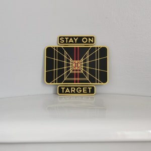 May include: A black and gold Star Wars themed sign with a grid pattern and red lines. The sign reads "Stay on Target".