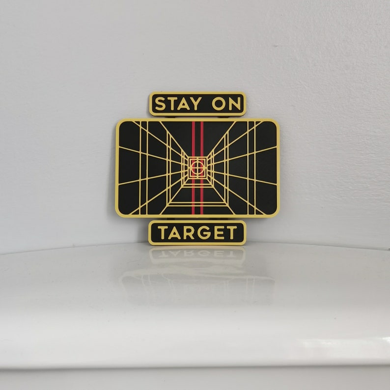 May include: A black and gold Star Wars themed sign with a grid pattern and red lines. The sign reads "Stay on Target".