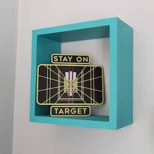 May include: A turquoise blue wooden frame with a black and gold graphic inside. The graphic is a stylized image of a target with the text "Stay on Target".