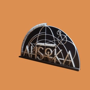 May include: A black and white acrylic sign with the text "Star Wars Ahsoka" in white letters. The sign is shaped like a half-circle and features a geometric design.