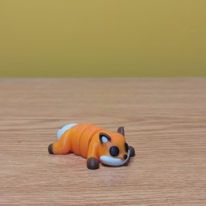 May include: A 3D printed orange and brown fox figurine with black eyes. The fox is lying on its belly with its tail curled up.