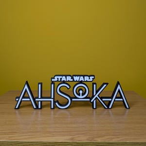 May include: A white 3D printed sign with the text "STAR WARS" above the text "AHsoka" in a stylised font. The sign is on a wooden surface.