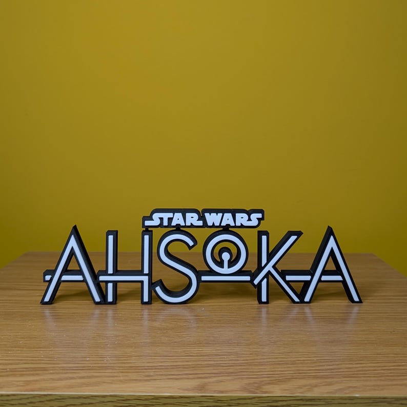 May include: A white 3D printed sign with the text "STAR WARS" above the text "AHsoka" in a stylised font. The sign is on a wooden surface.