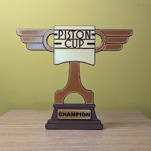 May include: A brown and gold Piston Cup trophy with wings, a piston-shaped base, and the words "Piston Cup" and "Champion" on it.