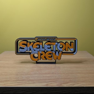 以下が含まれることがあります： 黒い背景にオレンジと青の文字で「STAR WARS SKELETON CREW」と書かれた3Dプリントされた看板です