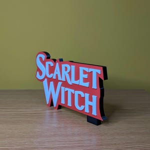 May include: A three-dimensional sign with the words "SCARLET WITCH" in white, set against a red background with a black border. The sign is displayed on a wooden surface with a yellow background.