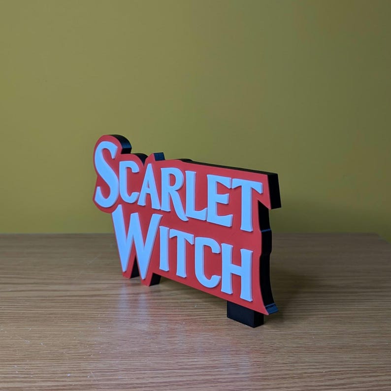 May include: A three-dimensional sign with the words "SCARLET WITCH" in white, set against a red background with a black border. The sign is displayed on a wooden surface with a yellow background.